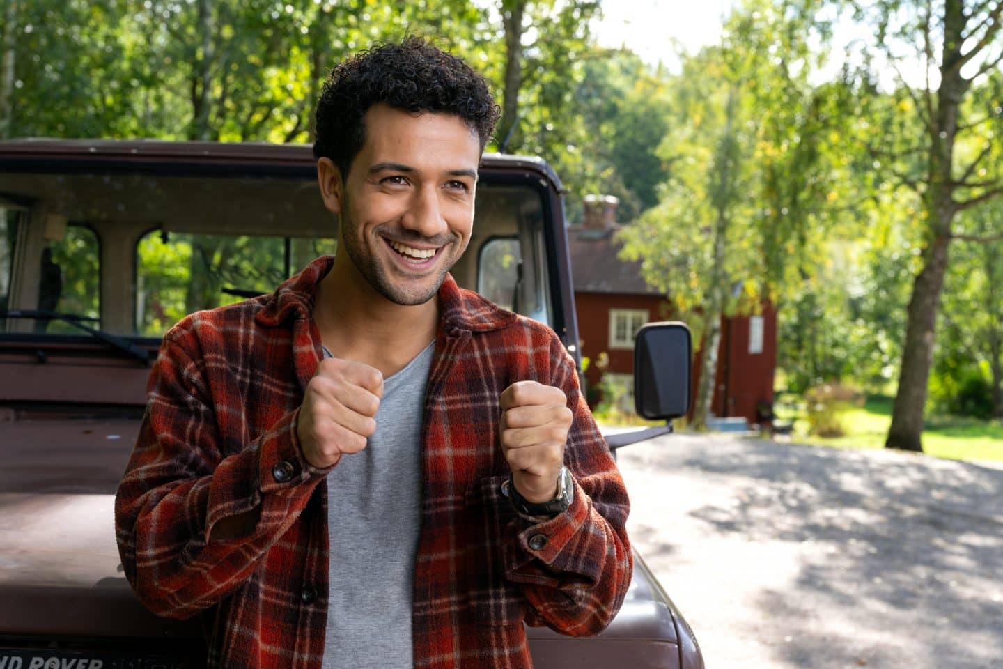 "Inga Lindström: Minnas Traum": Lucas (Rojan Juan Barani) steht an einer Straße vor einem Jeep. Er drückt beide Daumen und und blickt lächelnd geradeaus.
