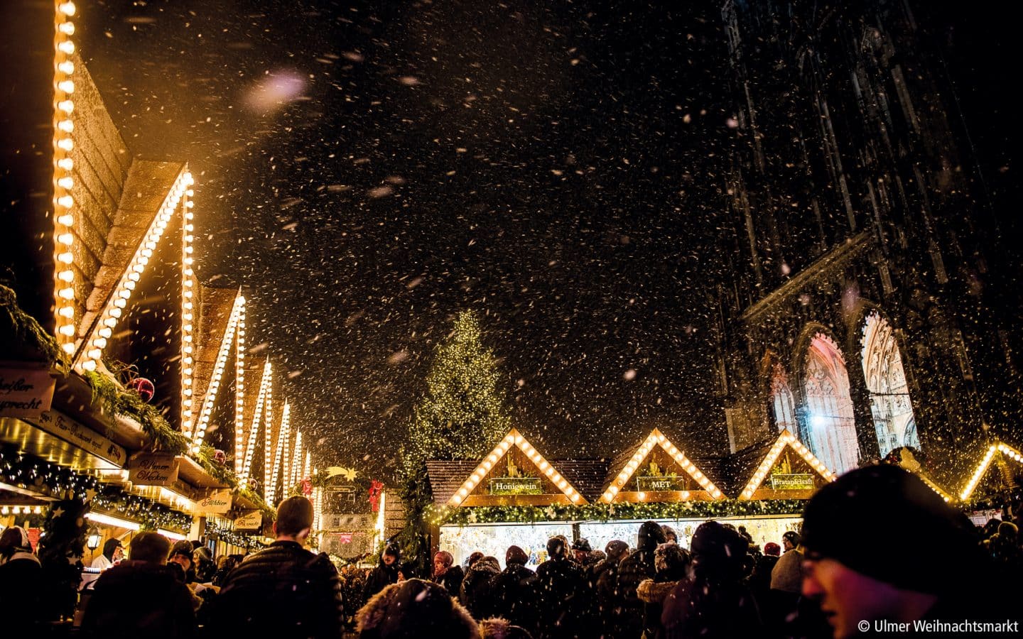 Verschneiter Weihnachtsmarkt mit beleuchteten Holzständen und Menschenmenge vor dem Ulmer Münster bei Nacht.