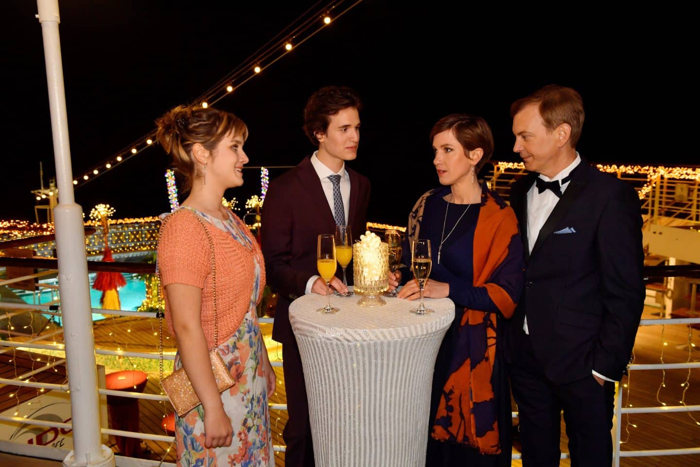 "Das Traumschiff: Afrika: Madikwe": Am Abend an Deck des Schiffs: Um einen Stehtisch herum stehen Lisa Sommer (Klara Kleinheins), Paul Wagner (Cooper Dillon), Katja Sommer (Isabell Horn) und Björn Wagner (Daniel Krauss) und unterhalten sich. Alle Vier tragen festliche Kleidung. Das Schiff ist mit vielen leuchtenden Lichterketten geschmückt.